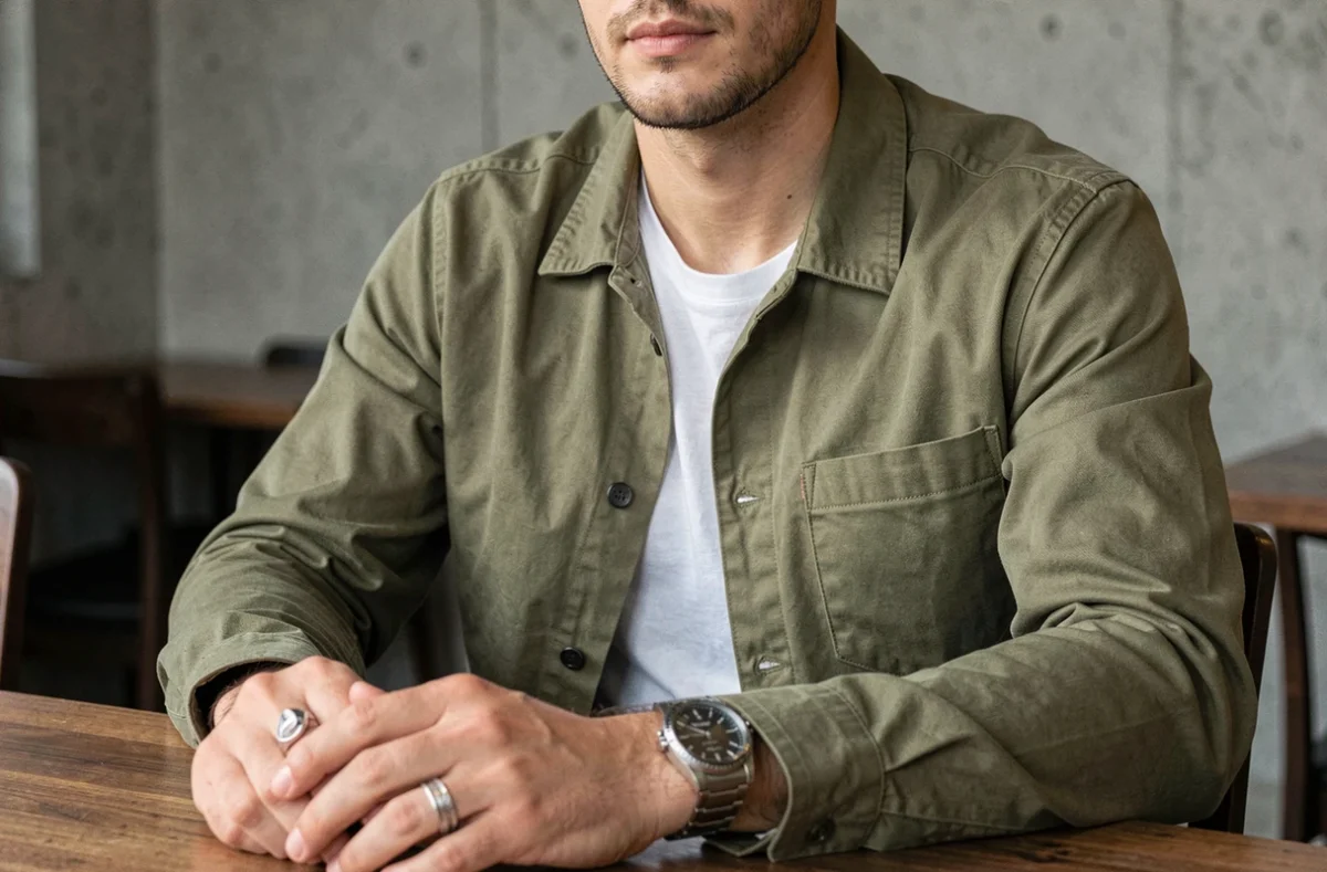 Homme en veste vert kaki avec montre et bague en argent dans un café minimaliste