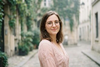 Jeune femme portant des lunettes tortoiseshell dorées et une robe midi rose romantique dans une cour parisienne en pierre